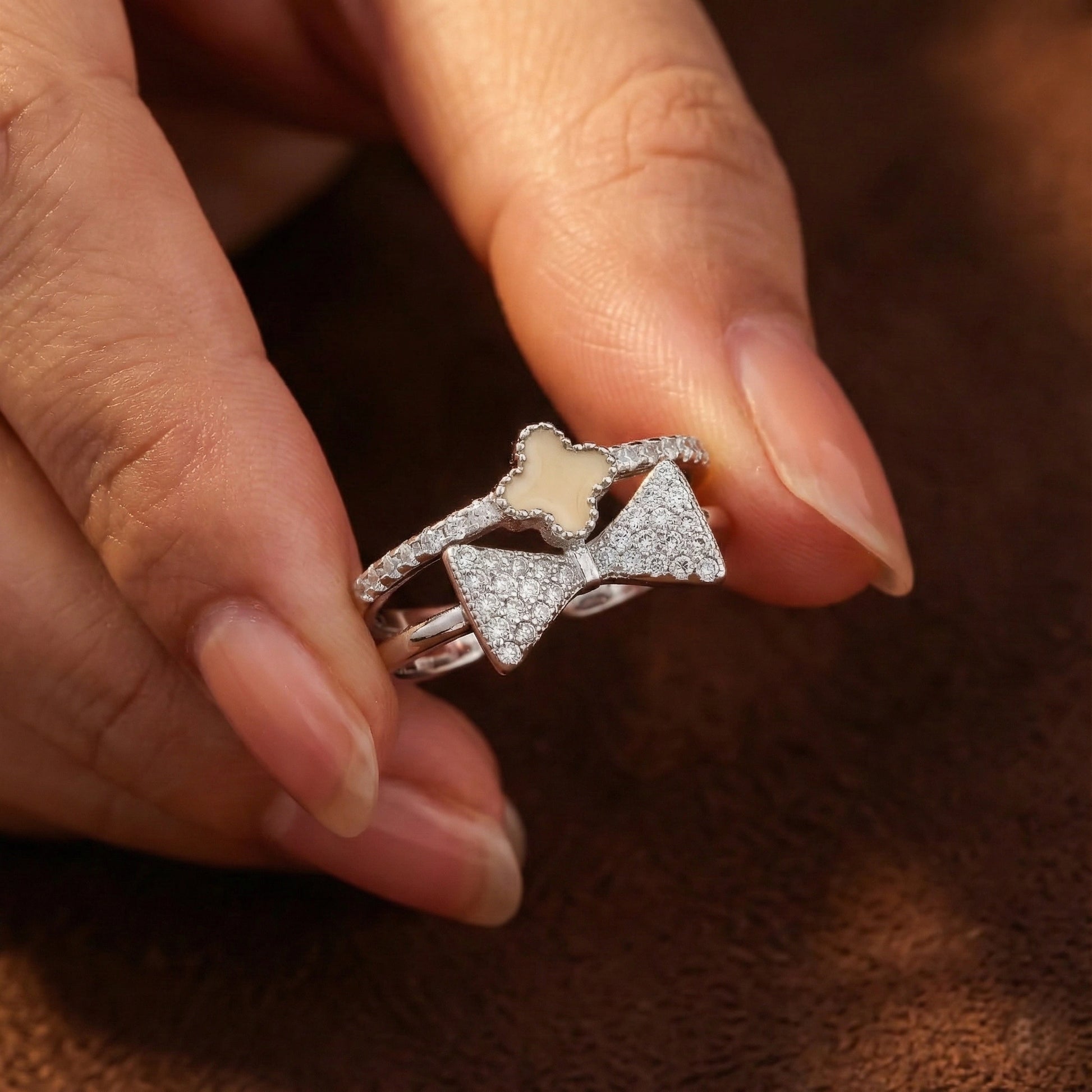 Silver ring with bow design held between fingers on a brown background