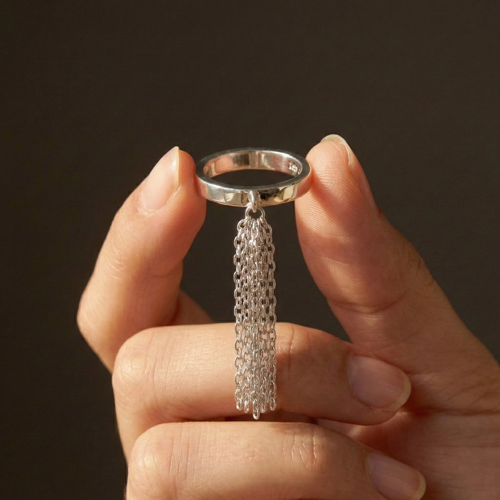 Silver ring with chain tassel held between fingers against a dark background