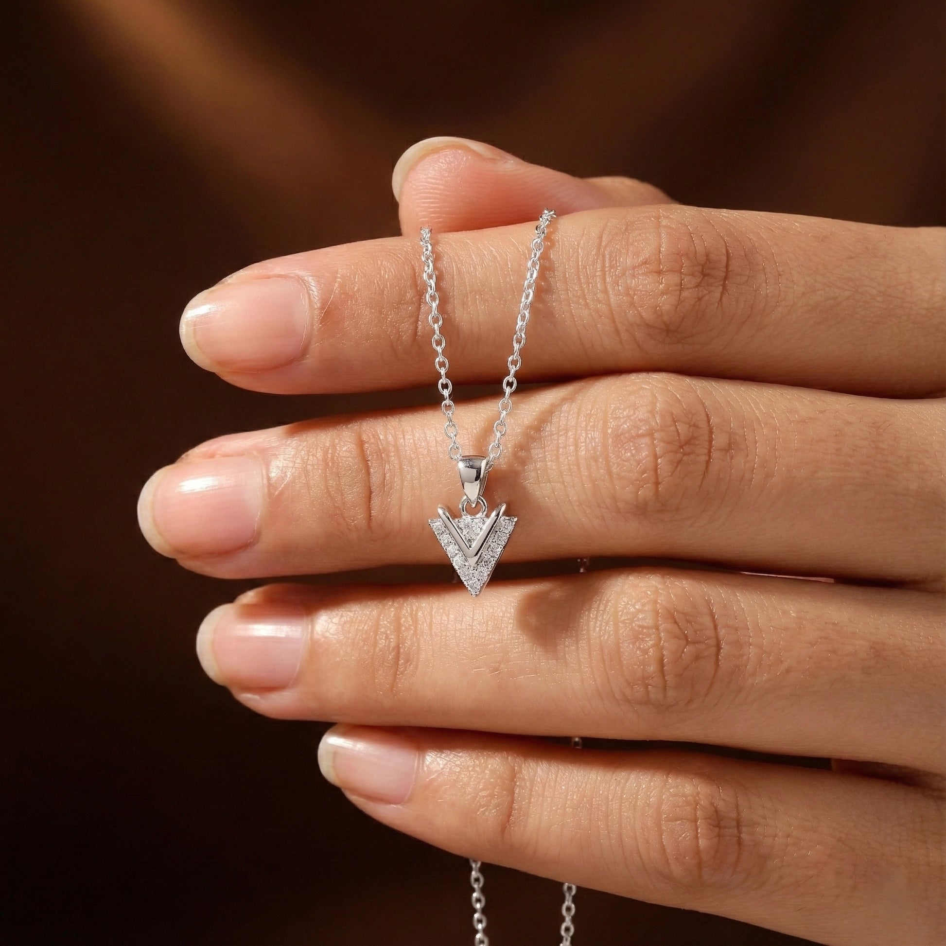 Silver necklace with a triangle pendant held between fingers against a brown background