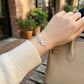 Hand wearing a silver bracelet with a bow design, holding a beige handbag outdoors.