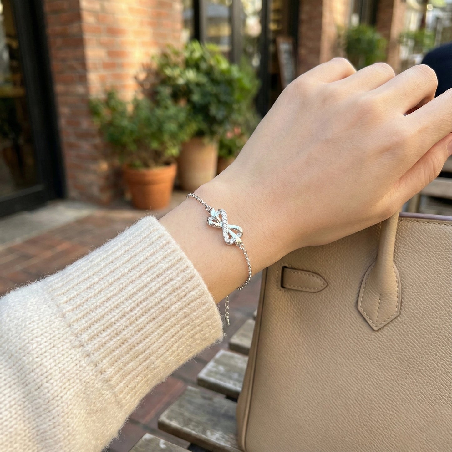 Hand wearing a silver bracelet with a bow design, holding a beige handbag outdoors.