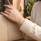 Hand holding a beige handbag with a 'love' bracelet on a blurred outdoor background