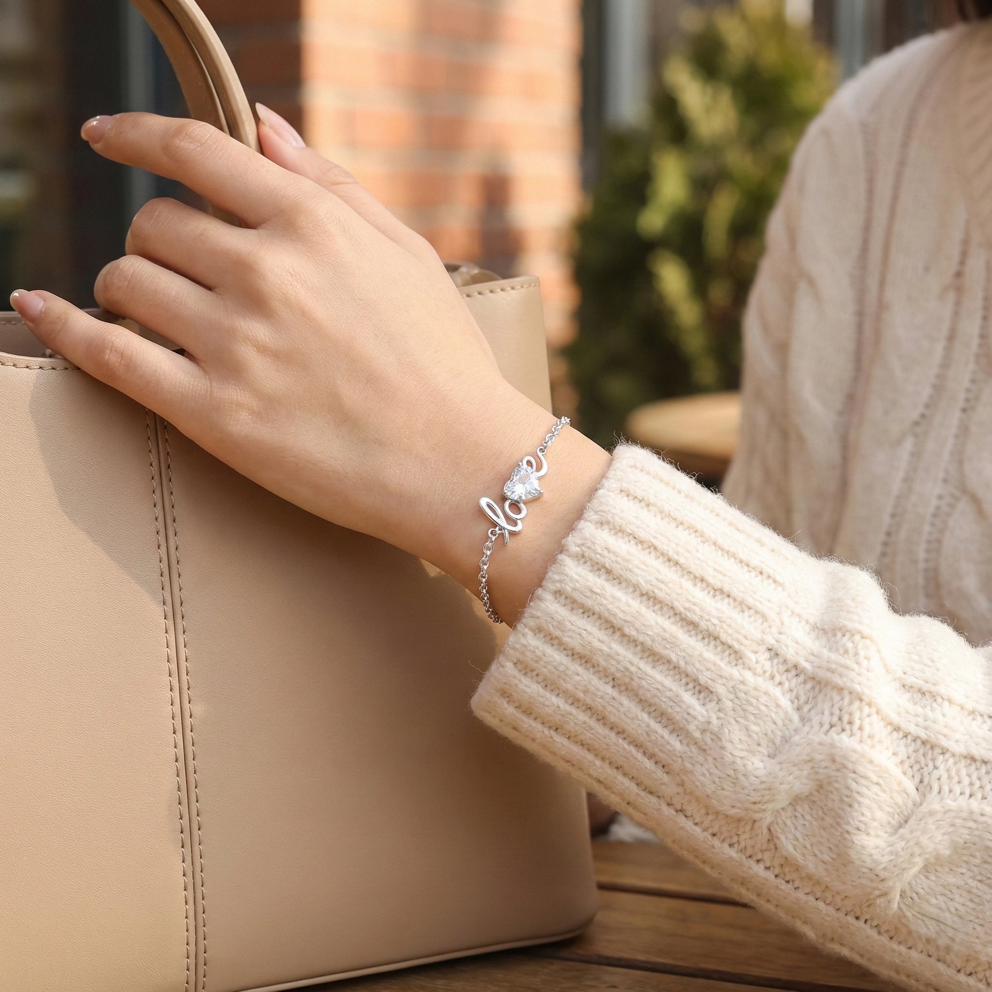 Hand holding a beige handbag with a 'love' bracelet on a blurred outdoor background