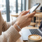 Person using a smartphone with a cup of coffee and laptop on a table in a cafe.