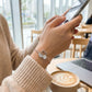 Person using a smartphone with a swan-shaped bracelet, surrounded by a coffee cup and laptop in a cafe setting.