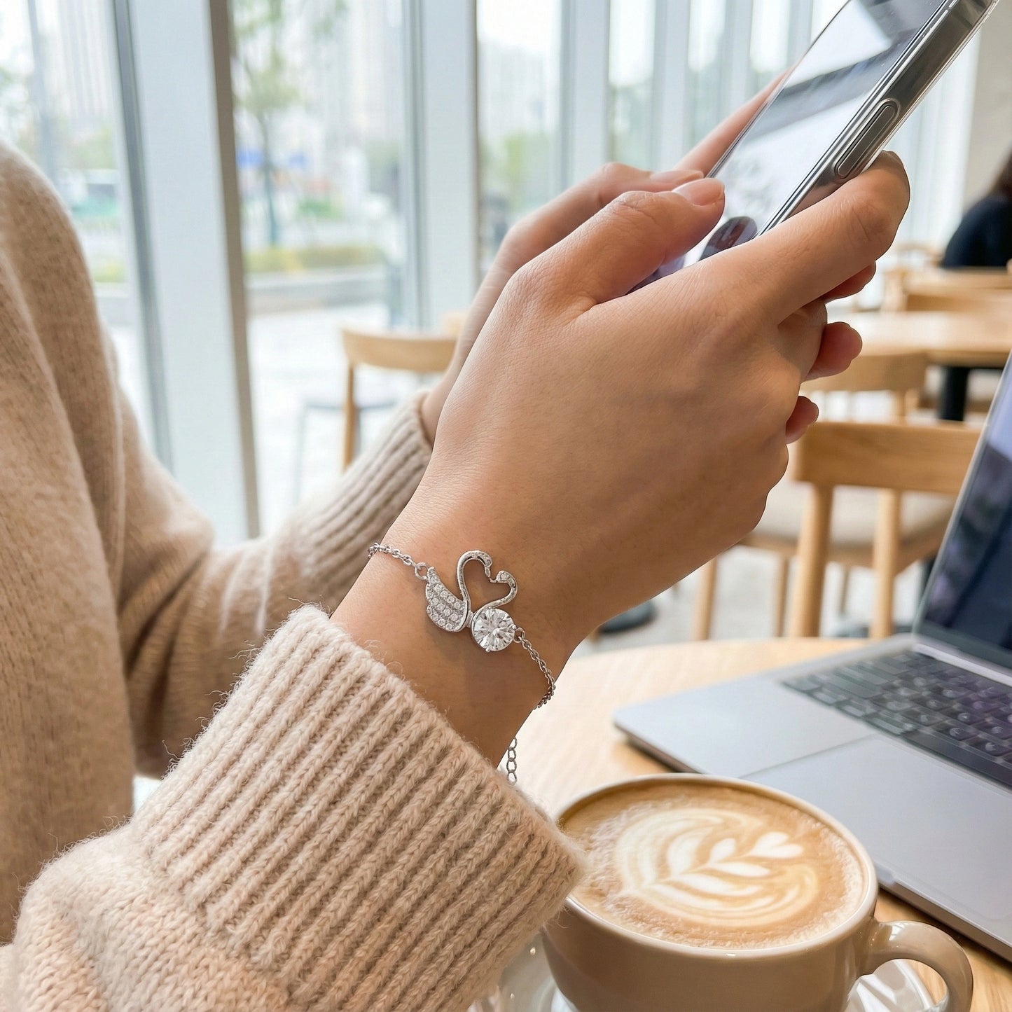 Person using a smartphone with a swan-shaped bracelet, surrounded by a coffee cup and laptop in a cafe setting.