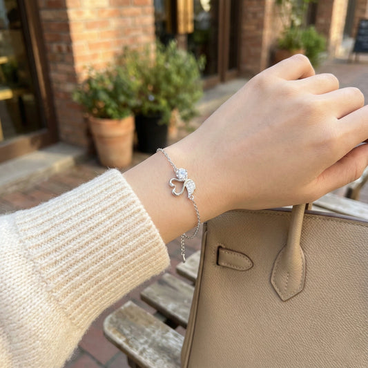 Hand wearing a silver bracelet with a heart charm, holding a beige handbag outdoors.