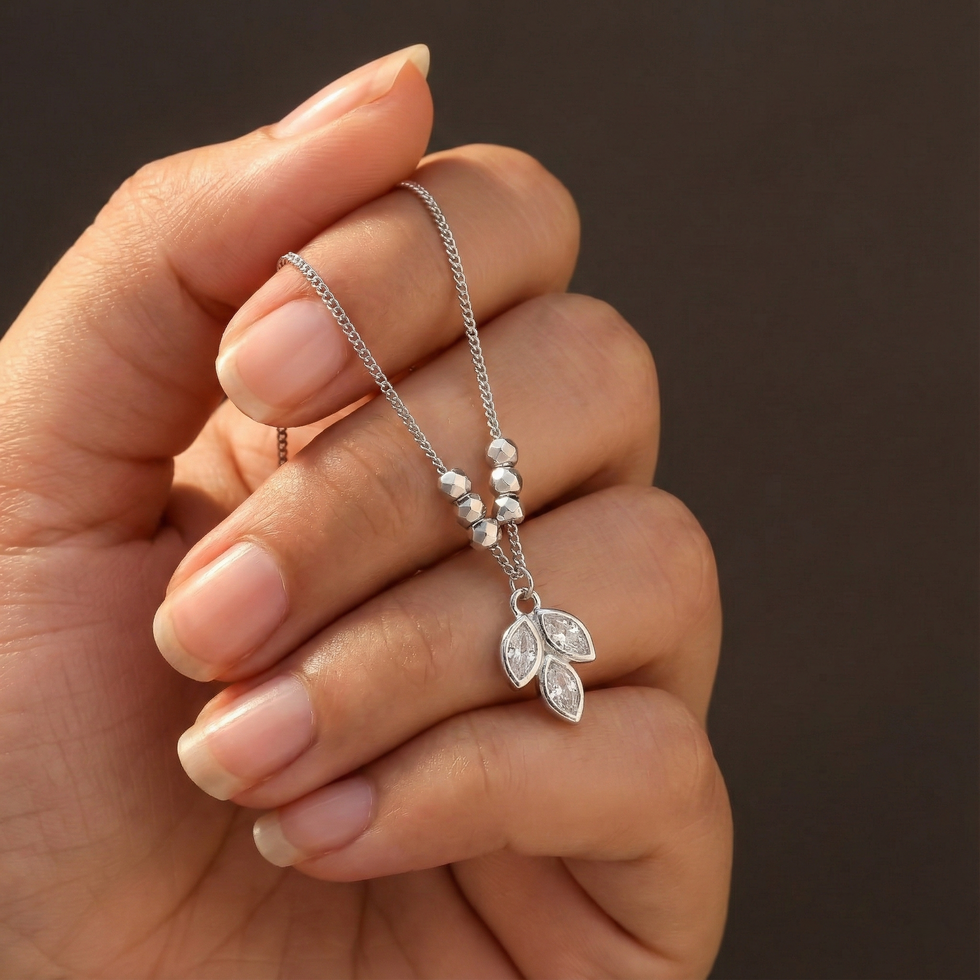 Hand holding a silver necklace with a leaf pendant against a brown background