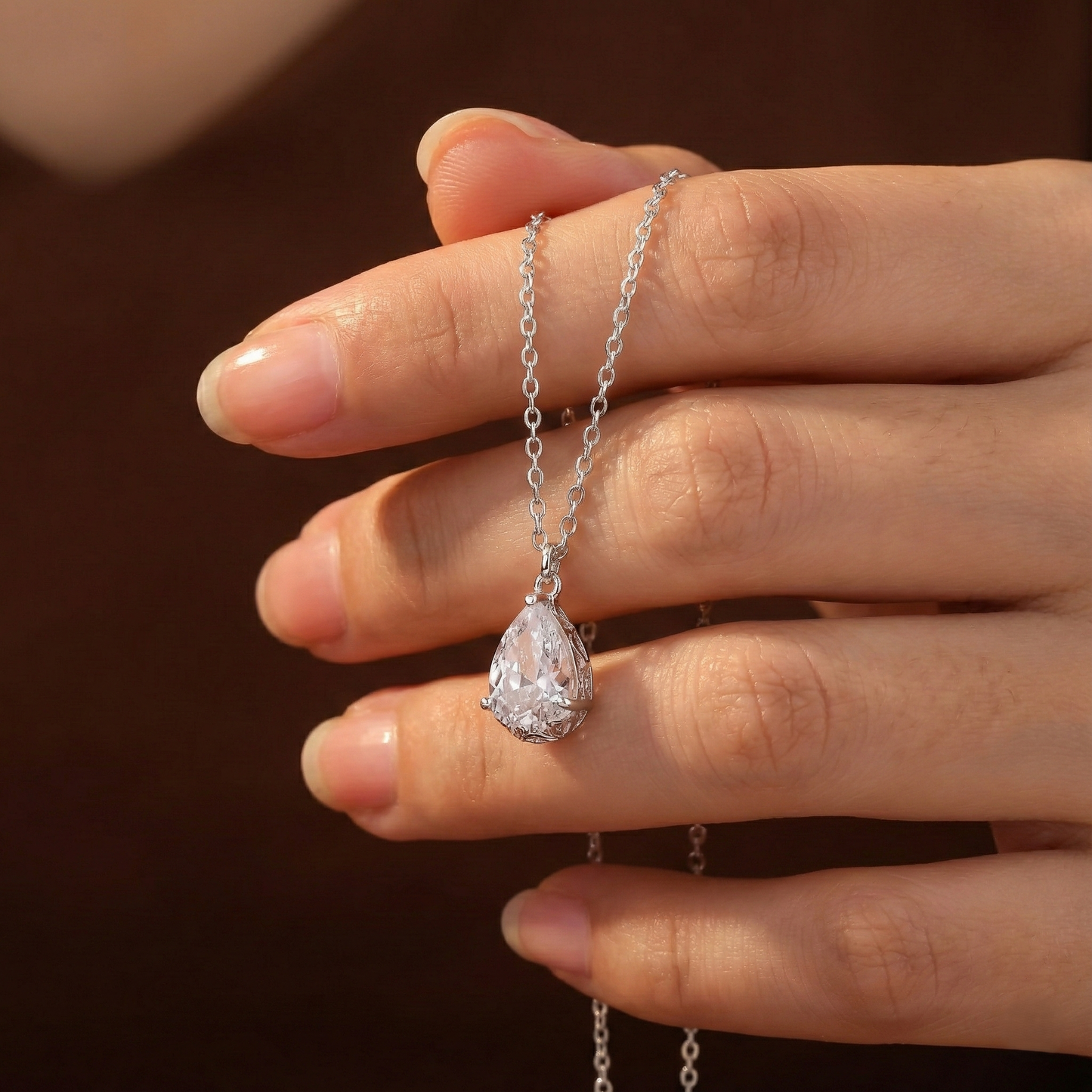 Hand holding a silver necklace with a teardrop-shaped diamond pendant against a dark background