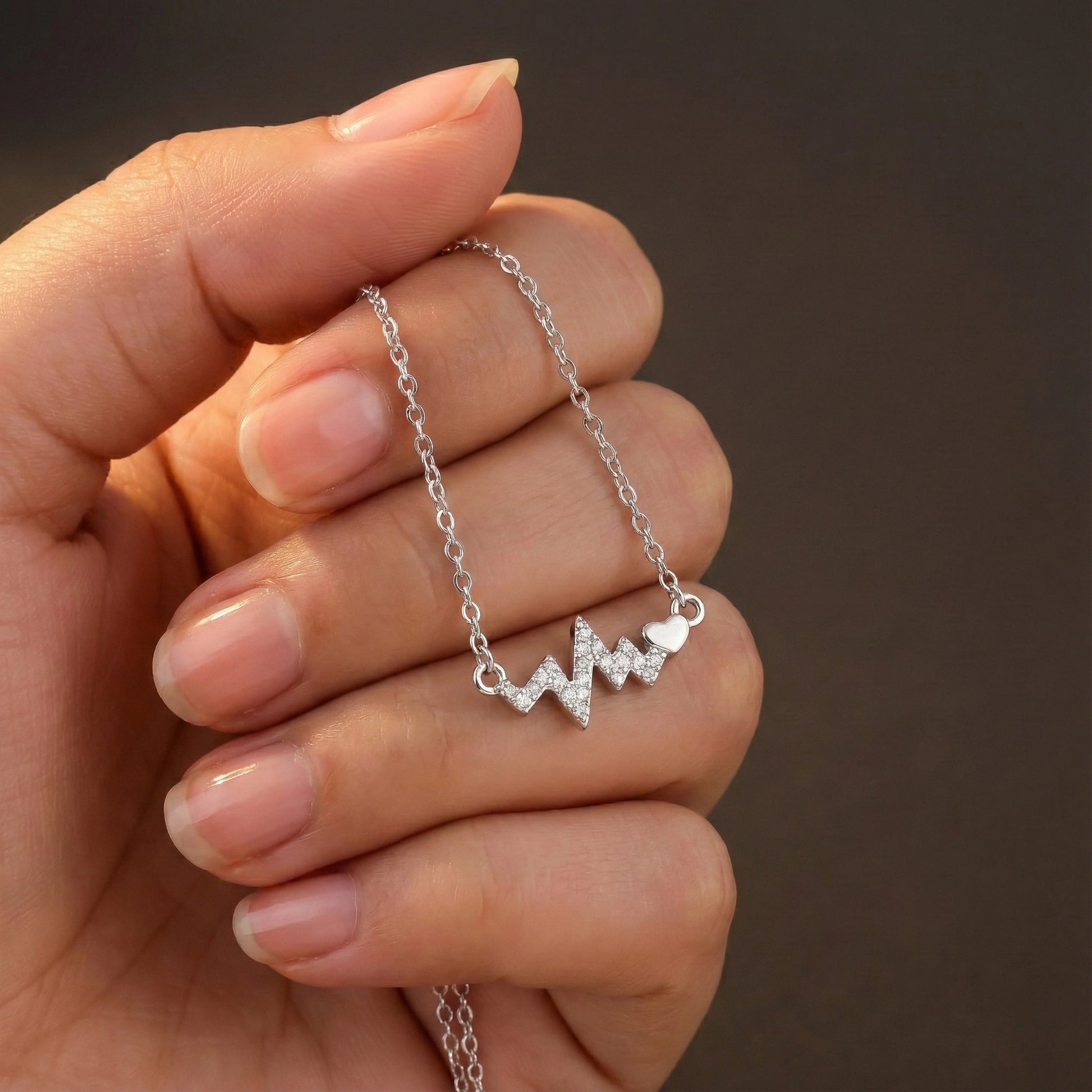 Hand holding a silver necklace with a heart and wave design against a brown background