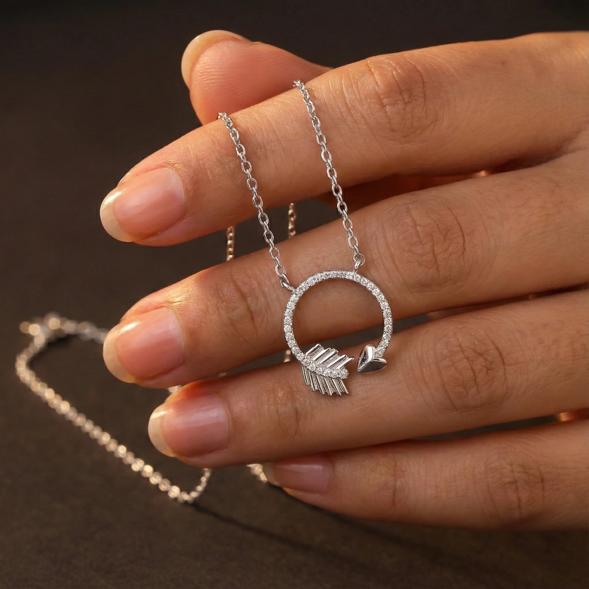 Silver necklace with a circular pendant featuring an arrow and heart held by a hand on a dark background