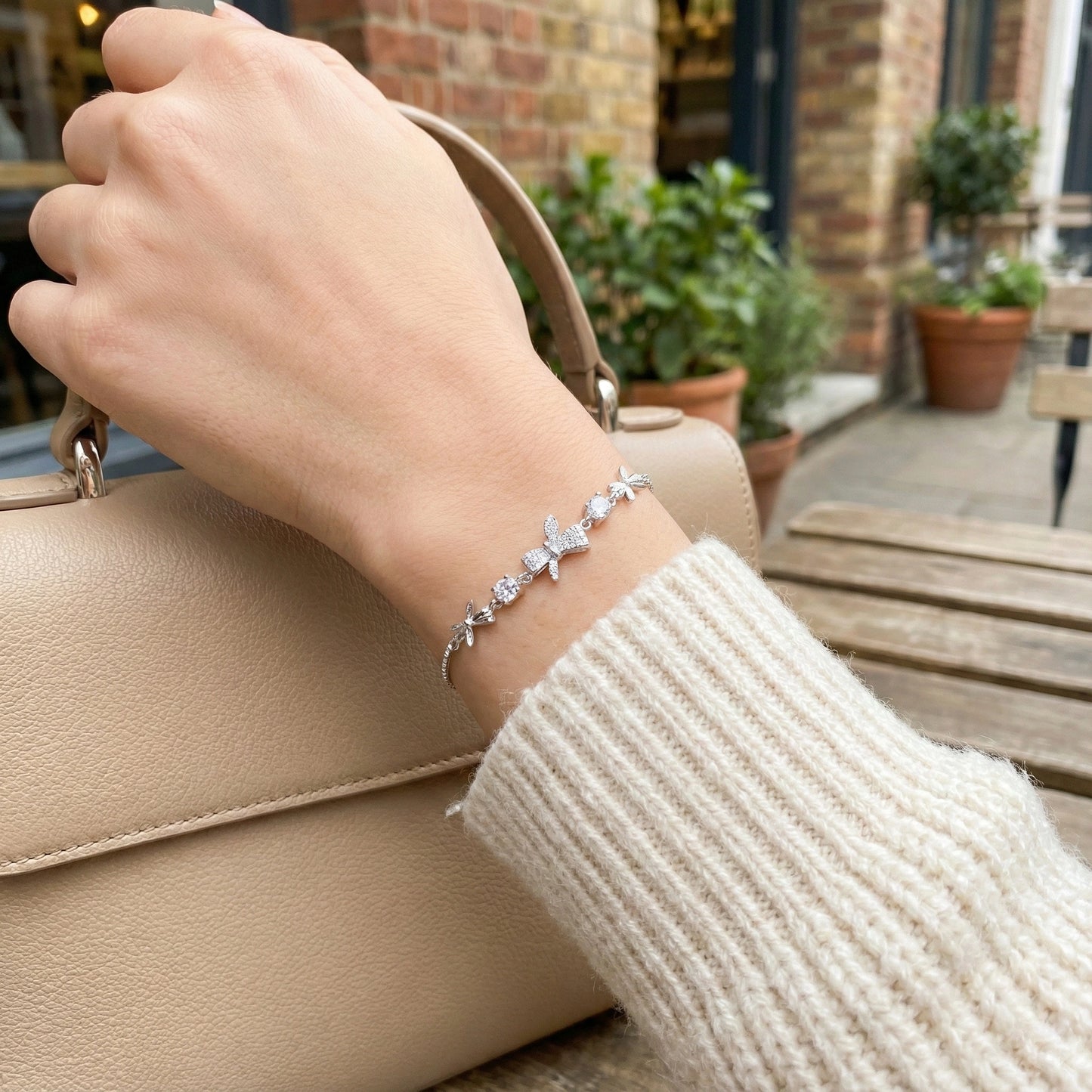 Hand holding a beige handbag with a bracelet on a blurred outdoor background