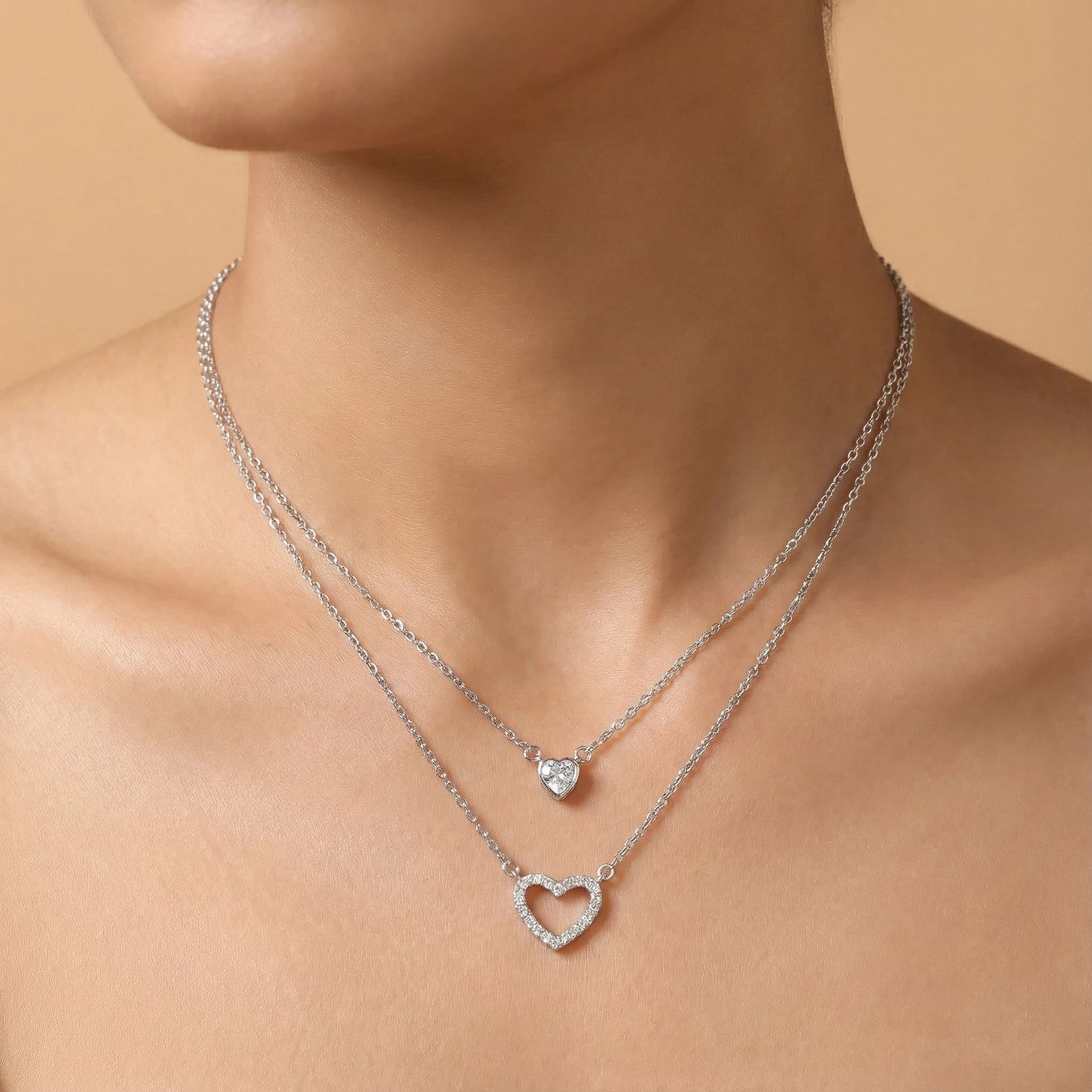 Silver necklaces with heart and round pendants on a person's neck against a beige background
