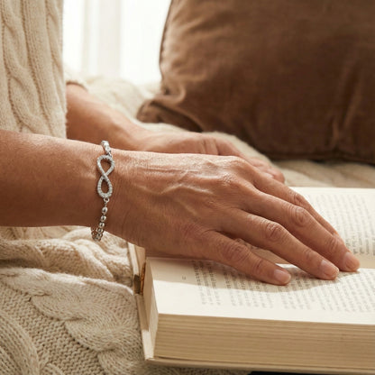 Person's hand with an infinity bracelet on a book, wearing a sweater and sitting on a couch.