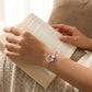 Person wearing a silver bracelet with a cat charm while holding an open book.