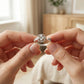 Two hands holding a silver ring with intricate design against a blurred indoor background