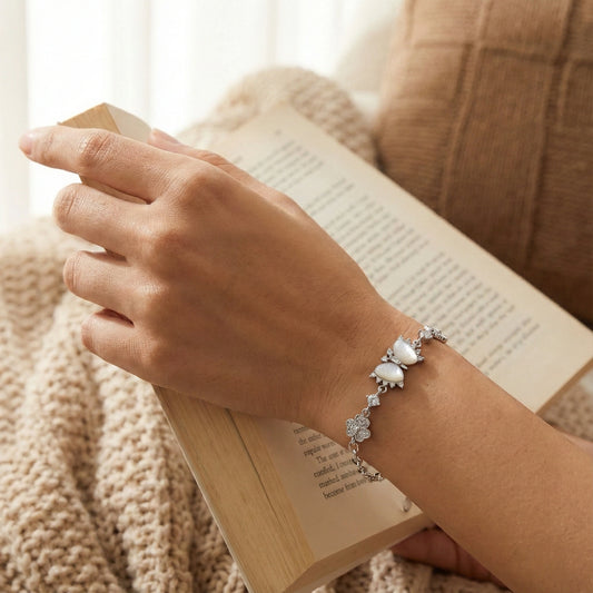 Hand wearing a silver bracelet with a heart charm, holding an open book against a soft, blurred background.