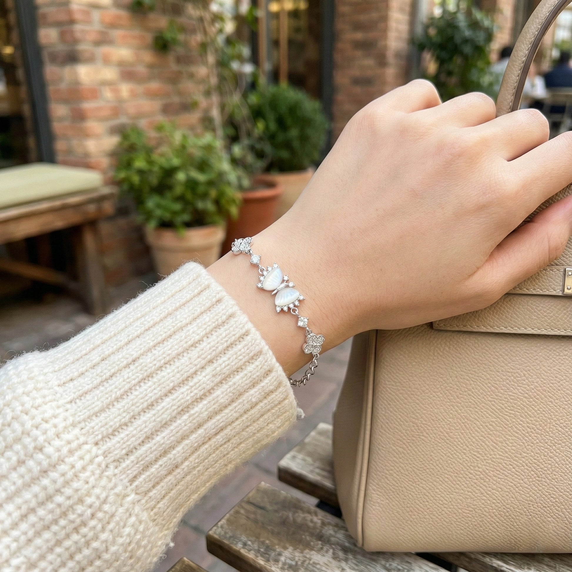 Hand wearing a silver bracelet with floral charms, holding a beige handbag outdoors.