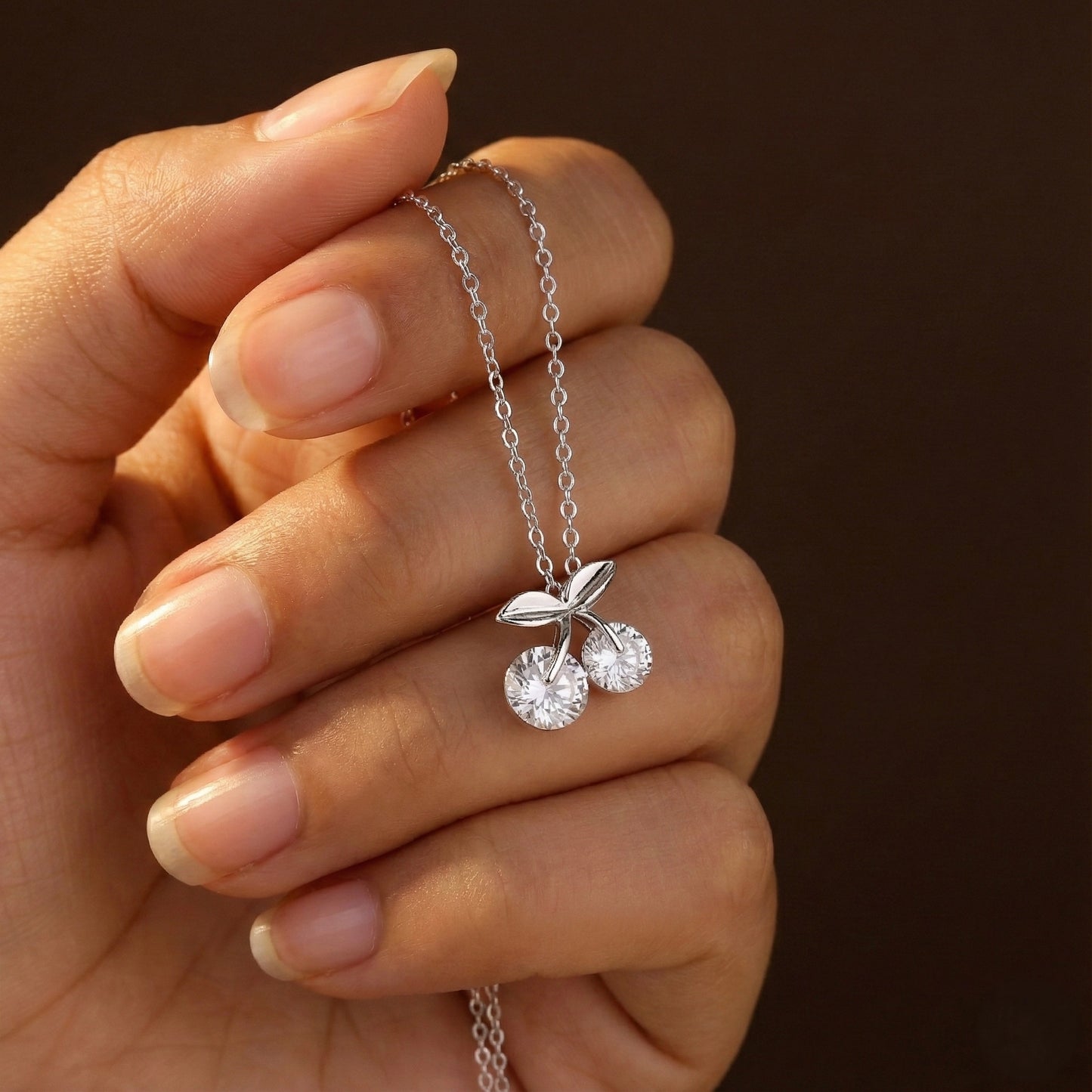 Hand holding a silver necklace with a cherry pendant against a dark background