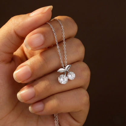 Hand holding a silver necklace with a cherry pendant against a dark background