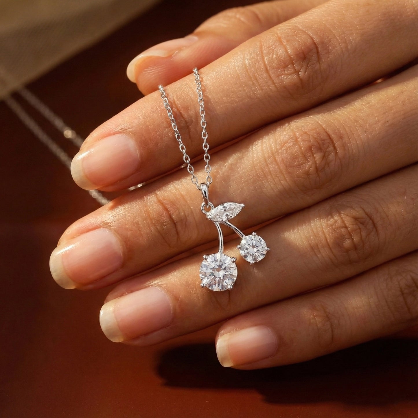 Silver cherry-shaped pendant necklace held by a hand against a blurred background