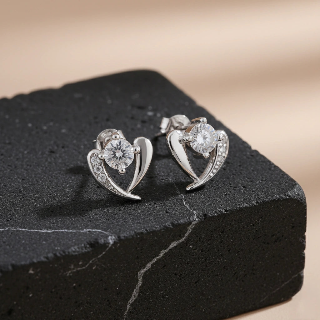 Heart-shaped silver earrings with clear stones on a black stone surface.