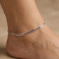 Close-up of a person's ankle with two thin anklets on a beige background