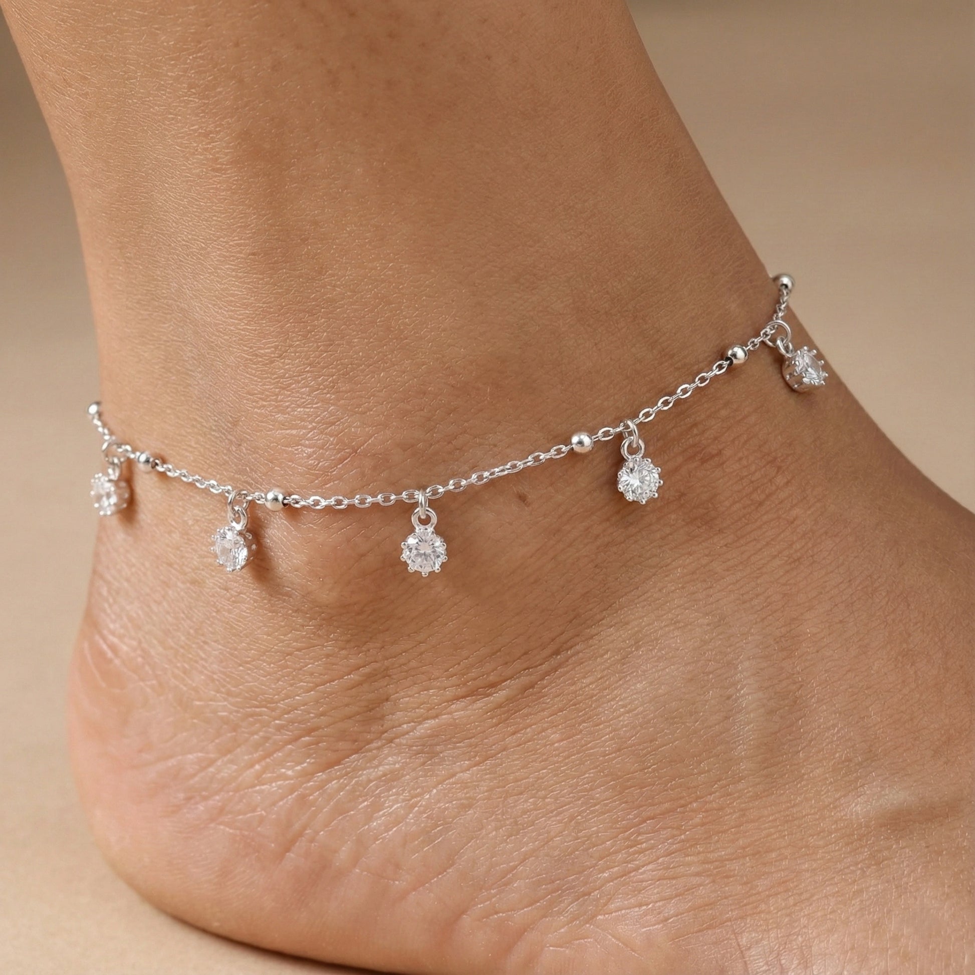 Silver anklet with small dangling charms on a foot against a beige background