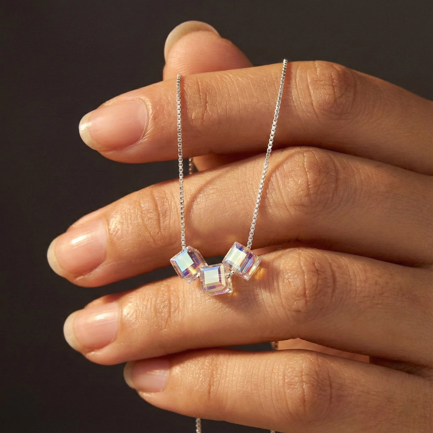 Hand holding a necklace with a cube-shaped pendant against a dark background