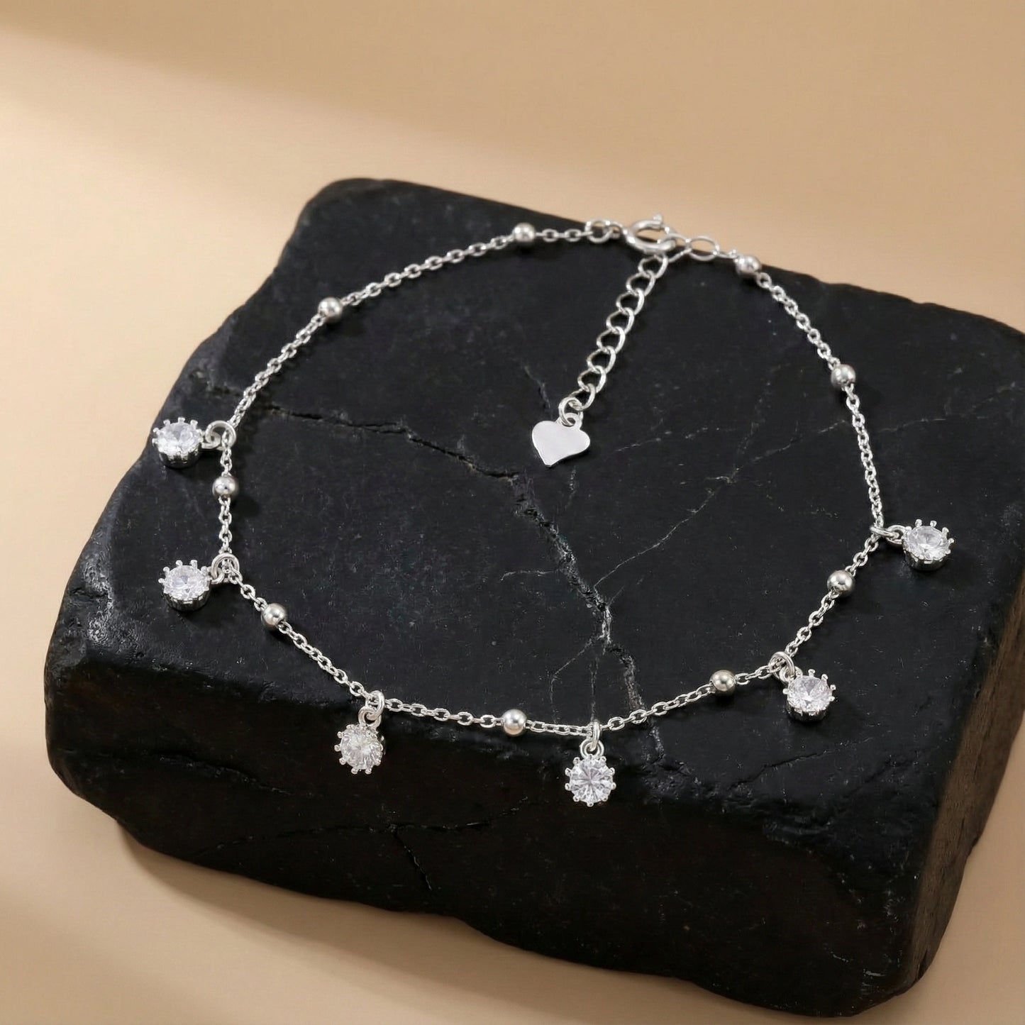 Silver anklet with heart and flower-shaped charms on a black stone surface