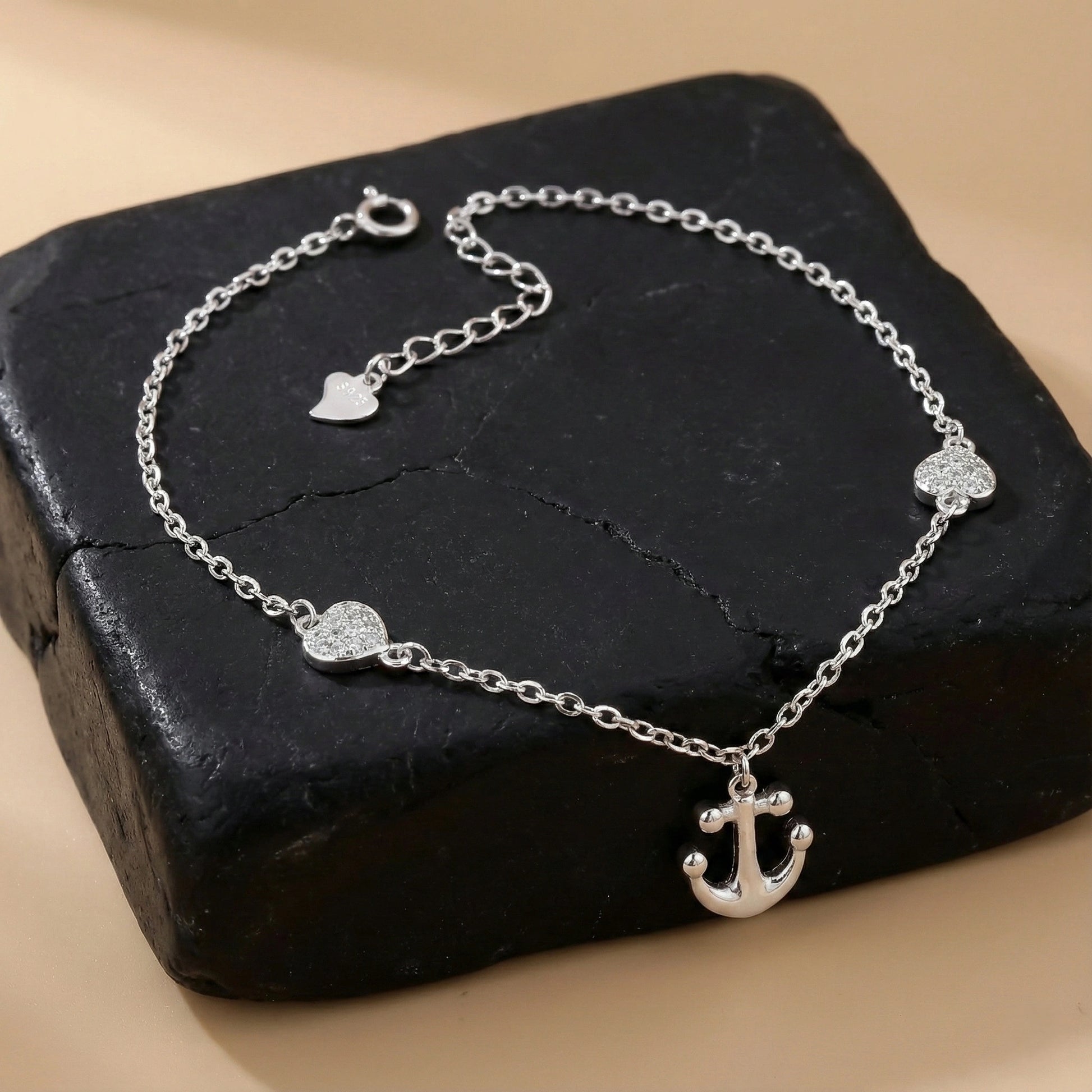Silver anklet with heart and anchor charms on a dark stone surface