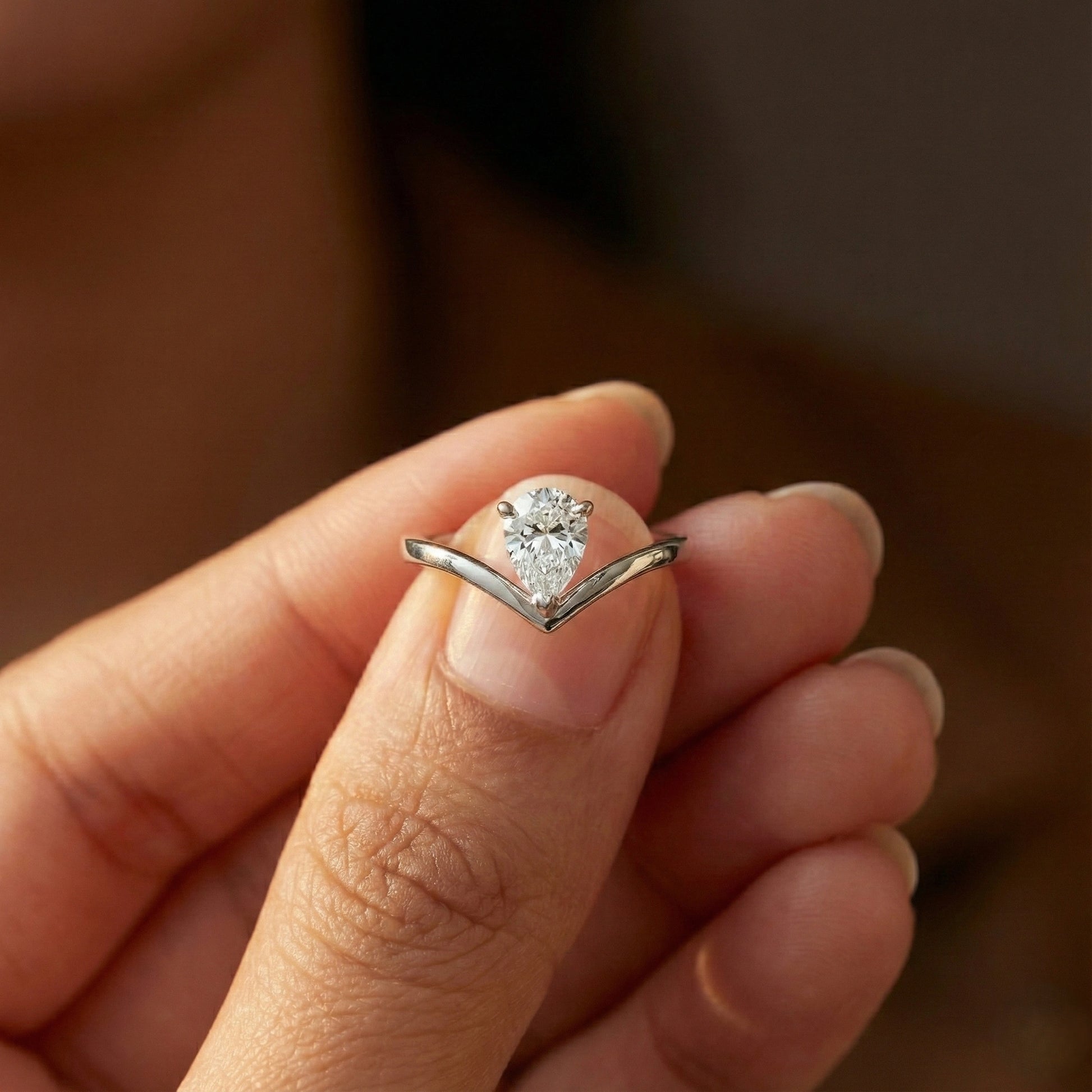 Close-up of a hand wearing a silver ring with a pear-shaped diamond on a blurred background