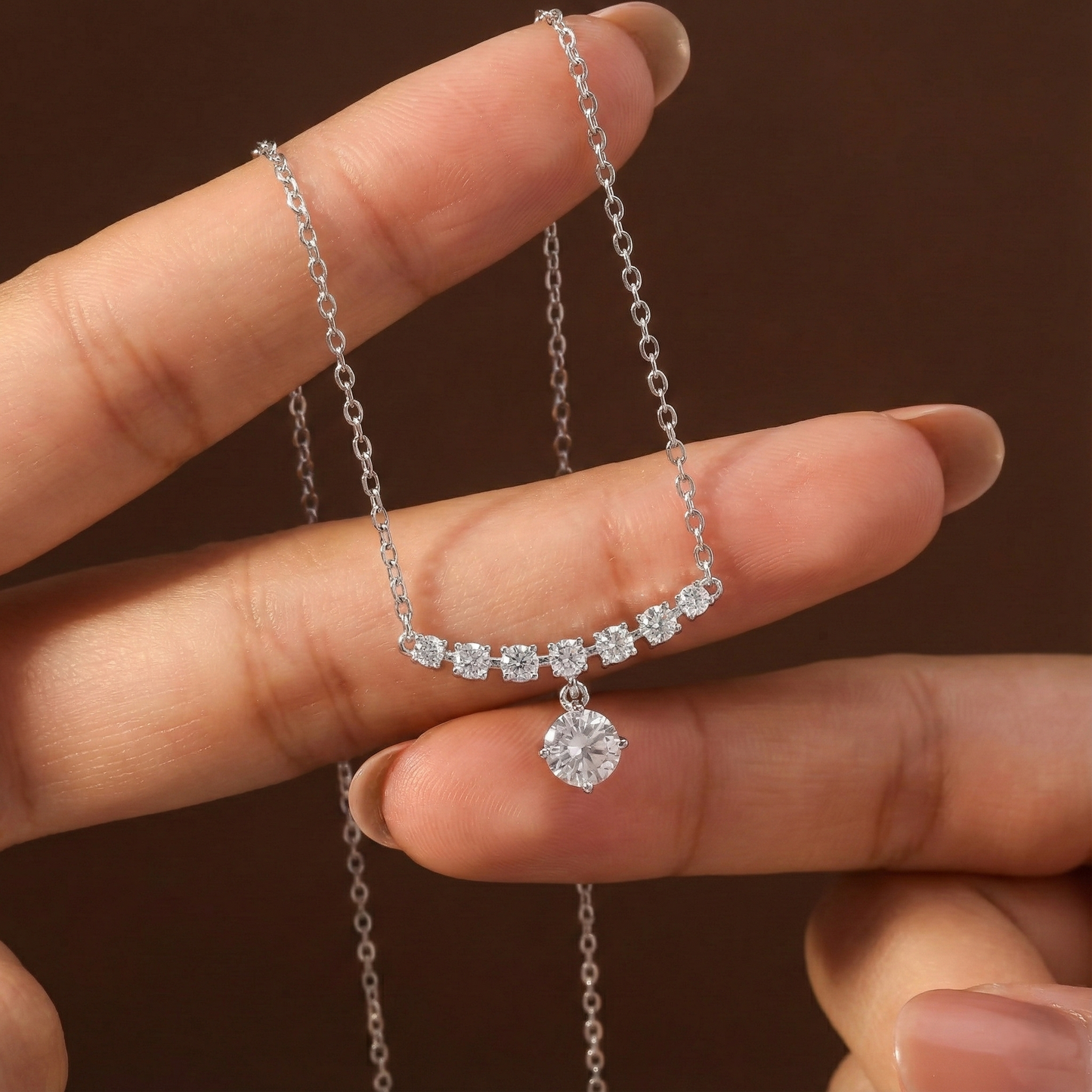 Silver necklace with a pendant held between fingers against a brown background