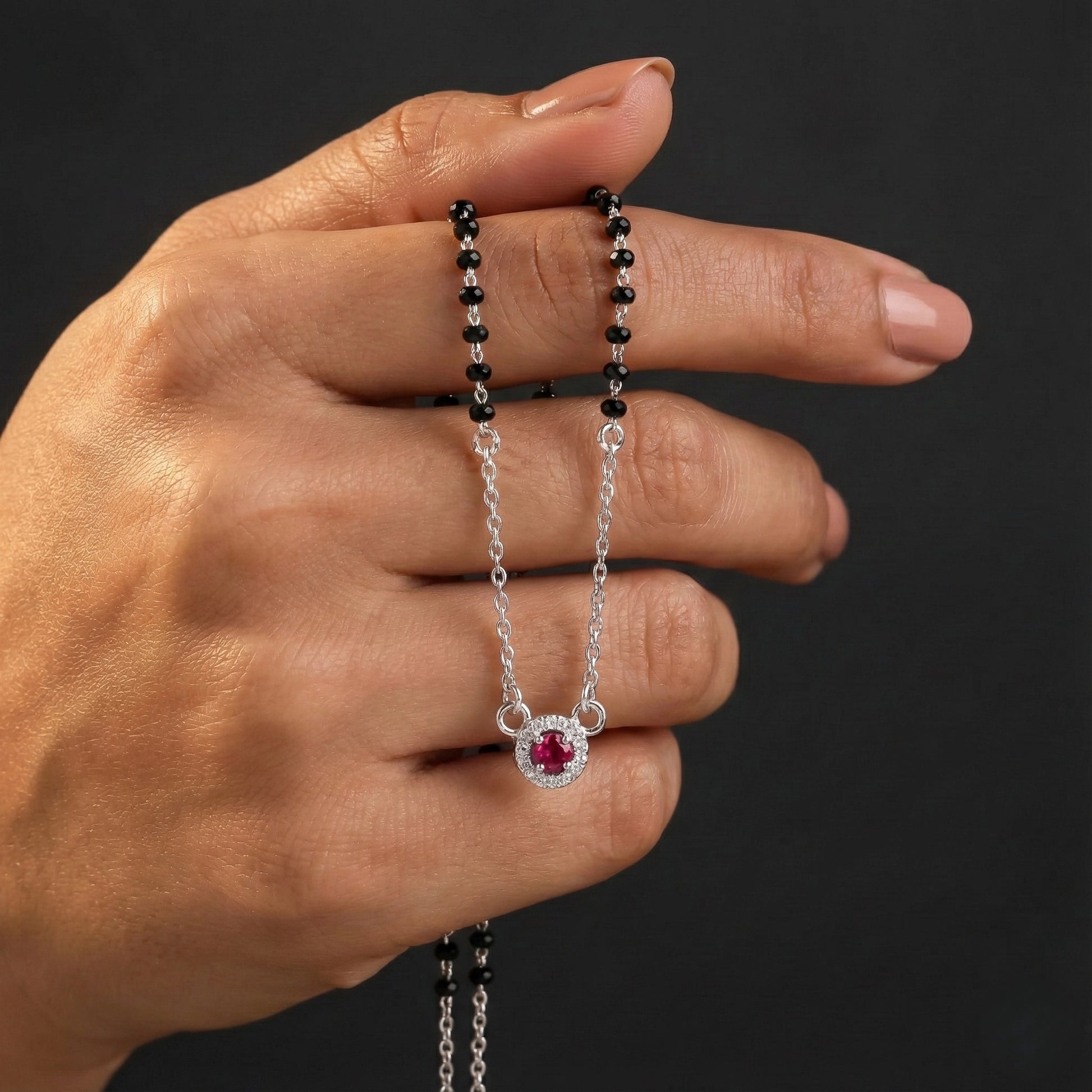 Hand holding a silver necklace with a red gemstone pendant against a dark background