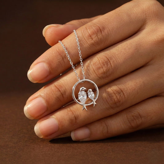 Silver necklace with a pendant of two birds held by a hand on a brown background