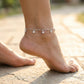Silver anklet with small charms on a foot standing on a stone surface.